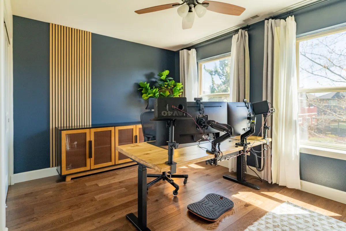 A focused view of a home office setup with a standing desk, dual wide monitors, a green plant, and large windows that let in abundant natural light. The mood is serene and productive, perfect for remote work or creative pursuits.
