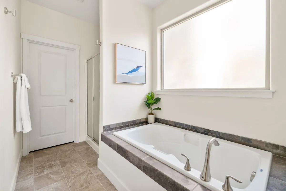 A tranquil ensuite bathroom featuring a soaking bathtub framed by a window, elegant artwork, and a potted plant. The abundance of natural light and thoughtful details create a spa-like ambiance ideal for relaxation and self-care routines.