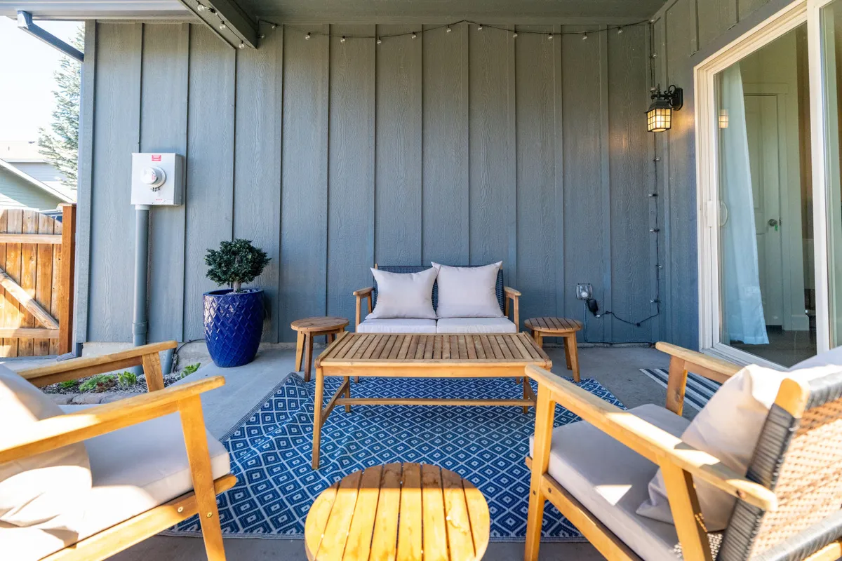 A stylish outdoor lounge setup with a sofa adorned with cushions, a wooden coffee table, a blue planter, and a decorative rug. The area looks comfortable and inviting, ideal for socializing or enjoying a coffee outdoors.