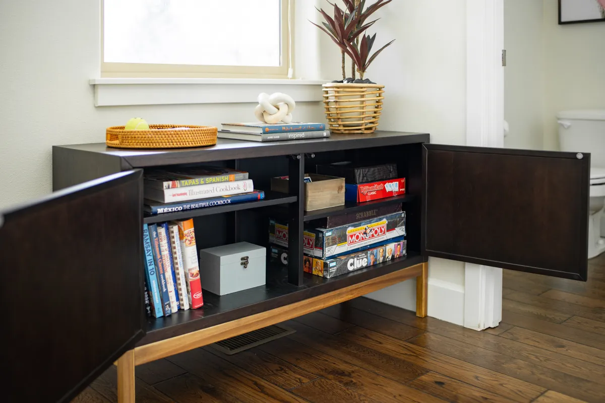 A welcoming corner with a wooden cabinet filled with books and board games, accompanied by an indoor plant. The arrangement invites guests to relax, read, or play games together, fostering a sense of connection and fun.