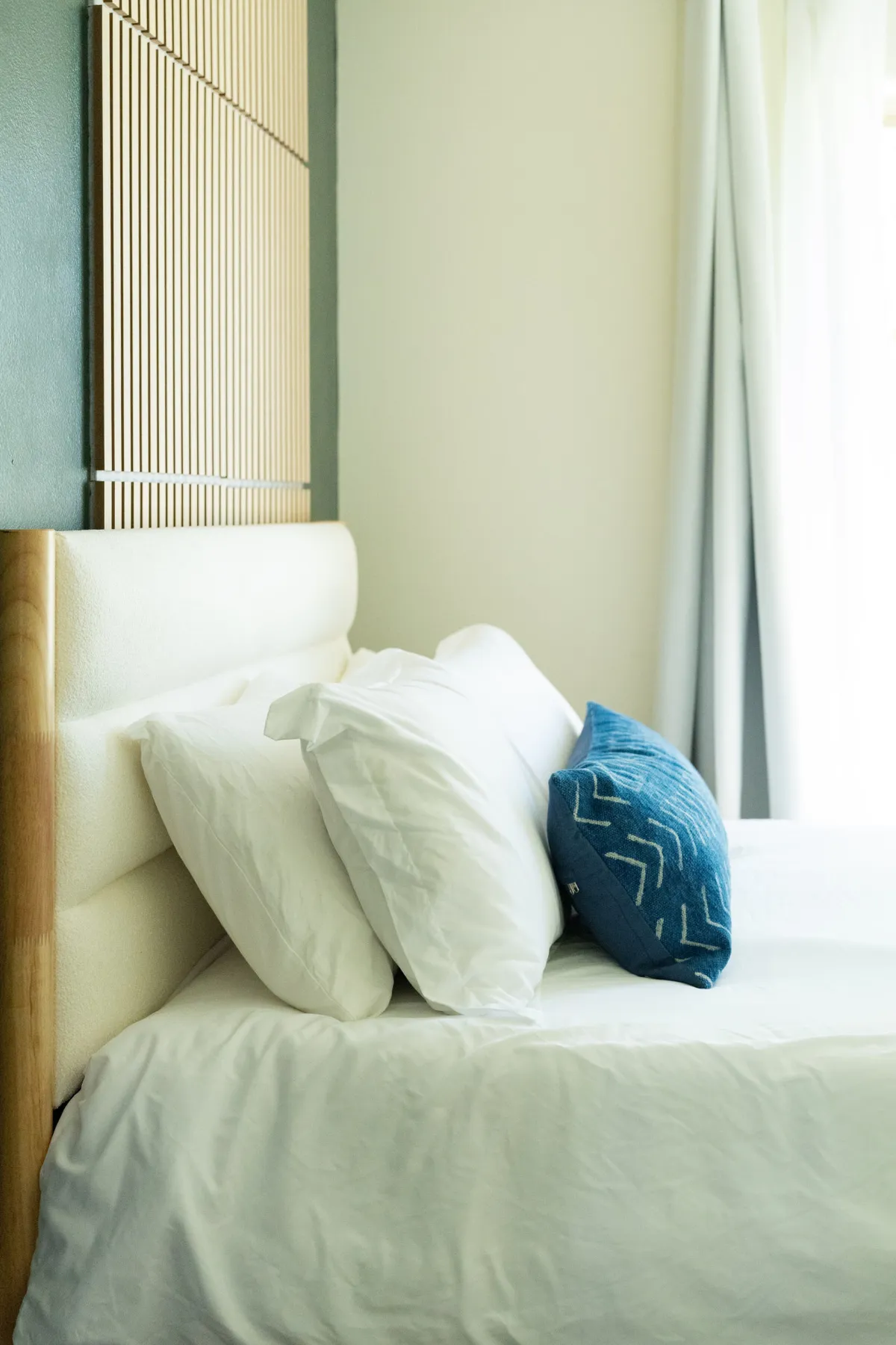 A serene bedroom with a bed featuring crisp white bedding, a decorative blue pillow, and a wooden headboard. Soft natural light filters into the room, enhancing the tranquil mood and inviting relaxation. The setup emphasizes both comfort and a calming atmosphere suitable for restful sleep.