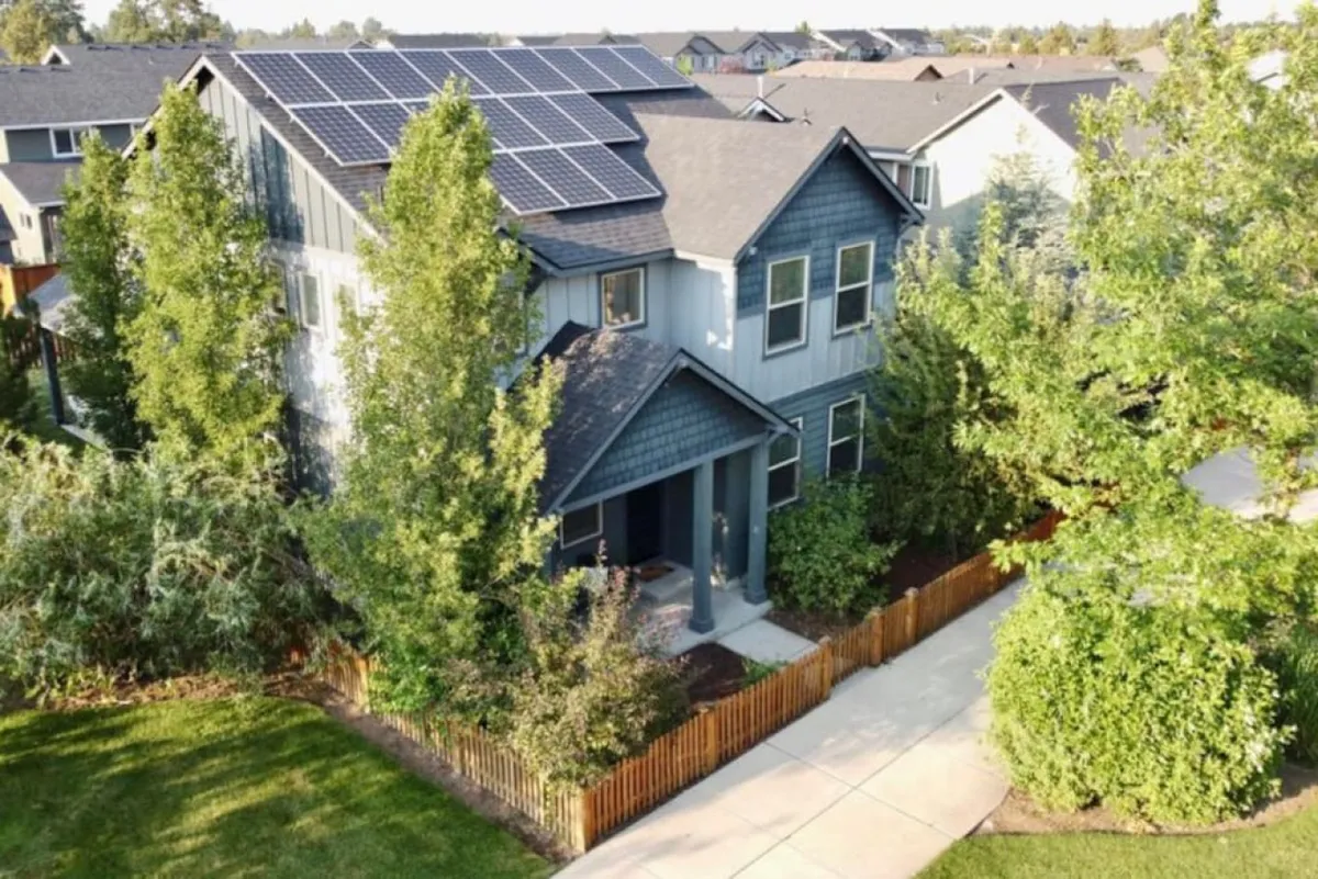 A welcoming exterior view of Bellflower House featuring solar panels, a cozy front porch, and a beautifully landscaped yard. The image captures the inviting atmosphere of the home, perfect for outdoor gatherings and neighborhood socializing.