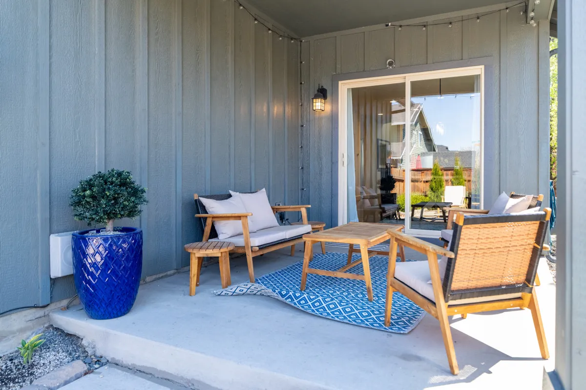 A cozy patio scene with wooden outdoor furniture arranged around a decorative rug, a blue planter adding a pop of color, and a sliding glass door connecting the inside to the out. String lights overhead give the space a warm glow, inviting relaxation whether the sun is rising or setting.
