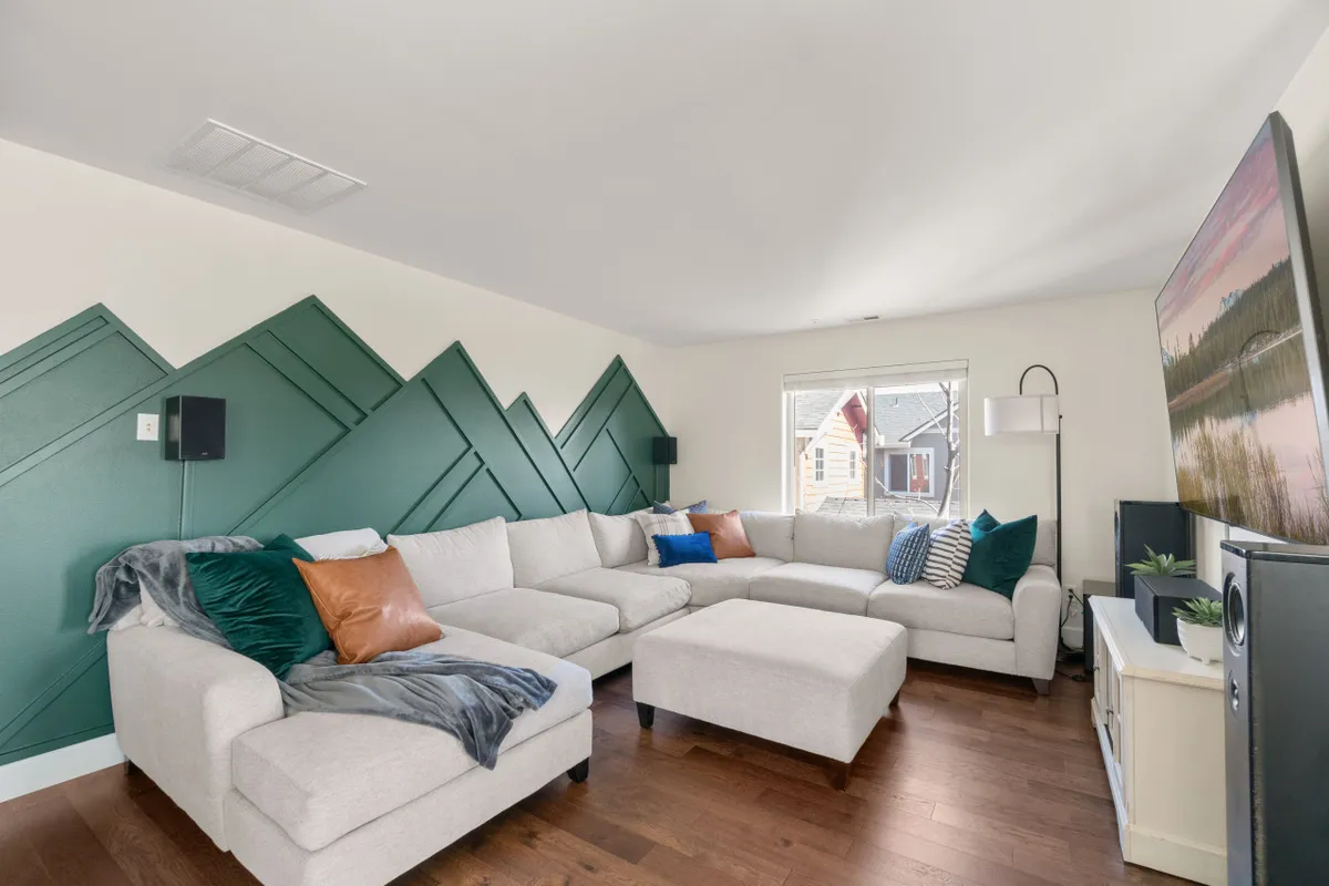 This interior image showcases a spacious living room with a large sectional sofa, a mountain-themed wall design, artwork, and plenty of natural light streaming in through the windows. The setup is perfect for socializing, relaxing, or enjoying a quiet morning coffee.