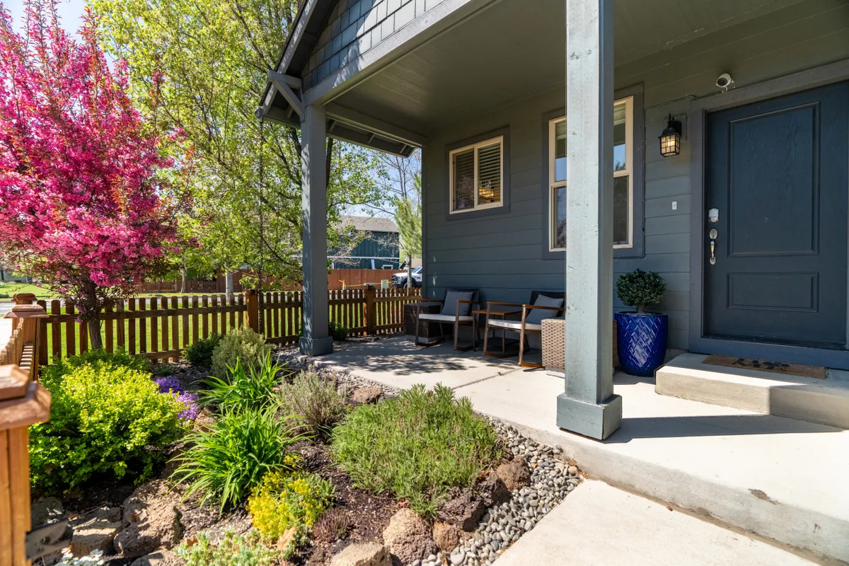 A charming exterior shot of Bellflower House with a pink flowering tree, lush green shrubs, cozy porch seating, and a welcoming front door. The scene is bright and cheerful, conveying a sense of arrival and home base for exploration.