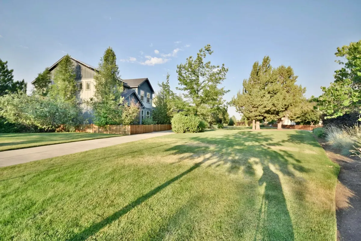 A lush green lawn stretches in front of the house, framed by mature trees and a pathway, suggesting ample space for outdoor gatherings, picnics, and play. The scene is tranquil and welcoming, with a strong emphasis on nature and community.