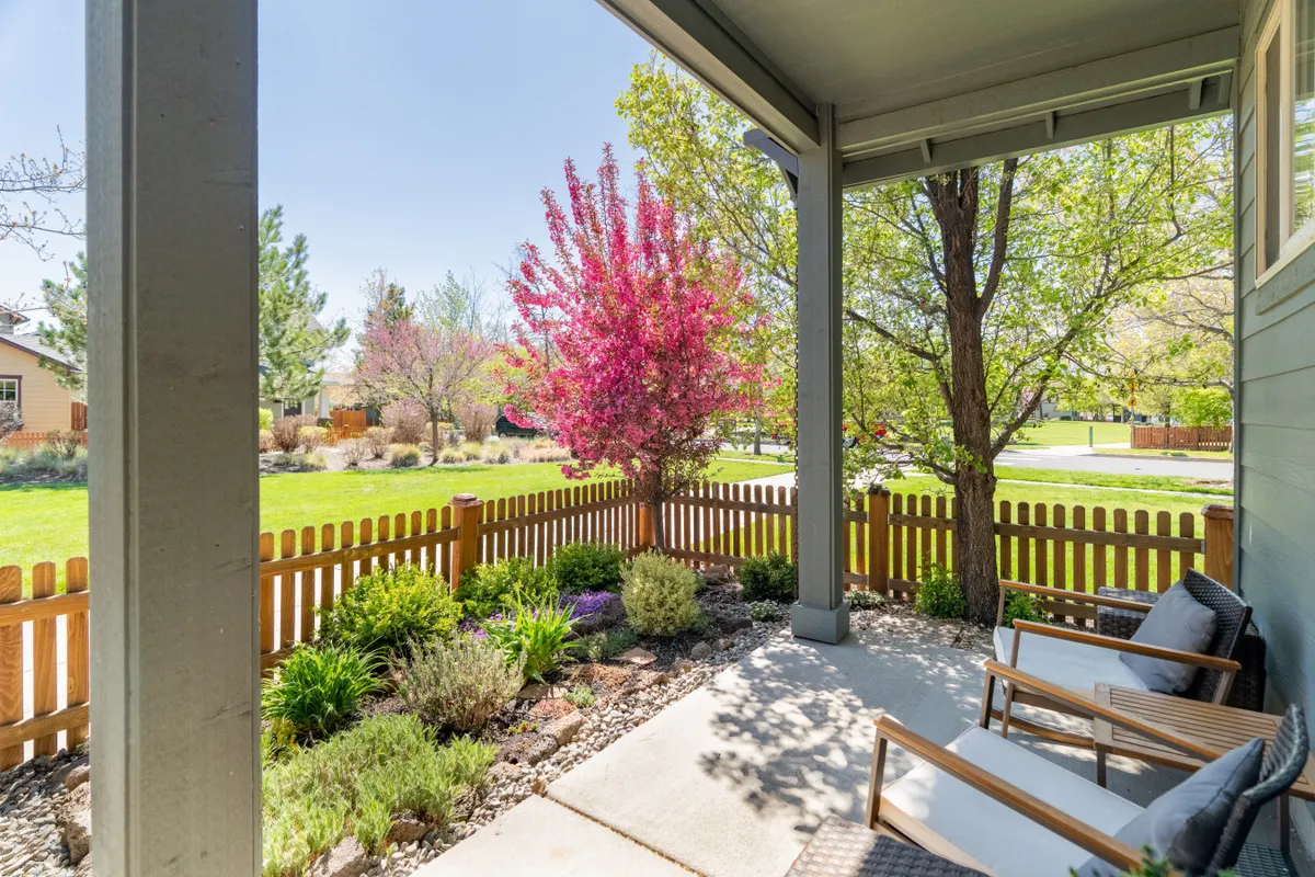 A peaceful backyard patio framed by a pink flowering tree, neatly trimmed garden, a wooden fence, and comfortable seating. The setting is inviting for both daytime relaxation and evening gatherings under twinkle lights.