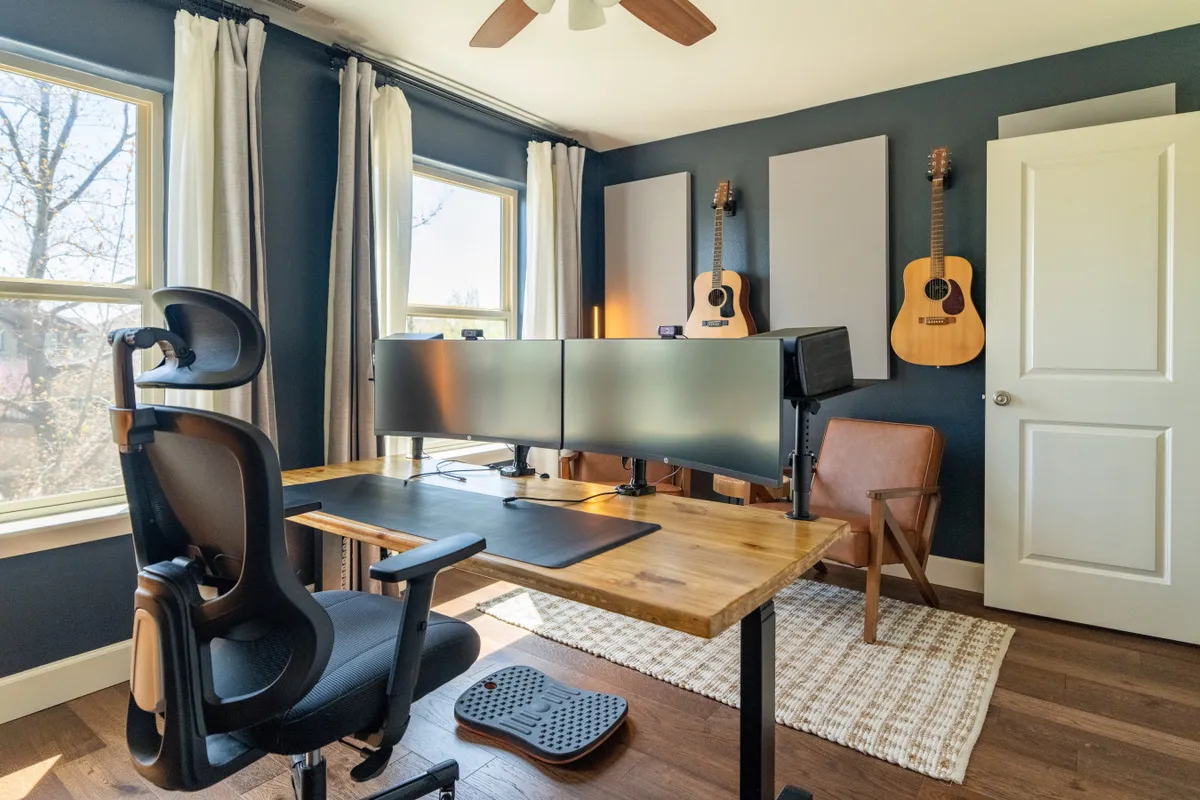 A creative workspace with dual computer monitors on a wooden desk and two acoustic guitars resting nearby. A comfortable chair and soft lighting complete the scene, hinting at musical gatherings and creative pursuits.