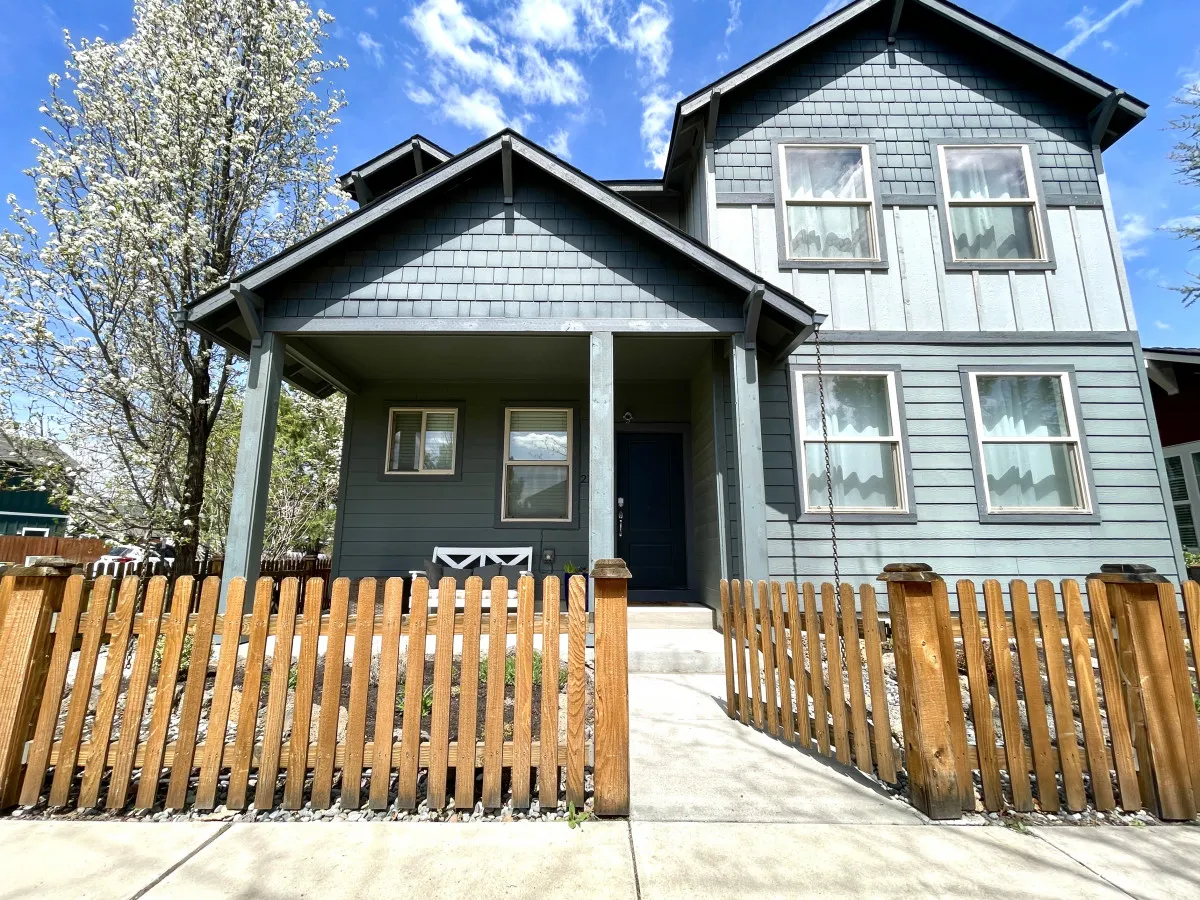 This exterior image showcases the front porch of Bellflower House, framed by a wooden fence and a vibrant flowering tree. The scene exudes a welcoming atmosphere, with sunlight casting a cheerful glow over the entry. It subtly hints at the kind of friendly, neighborly environment mentioned in the section.