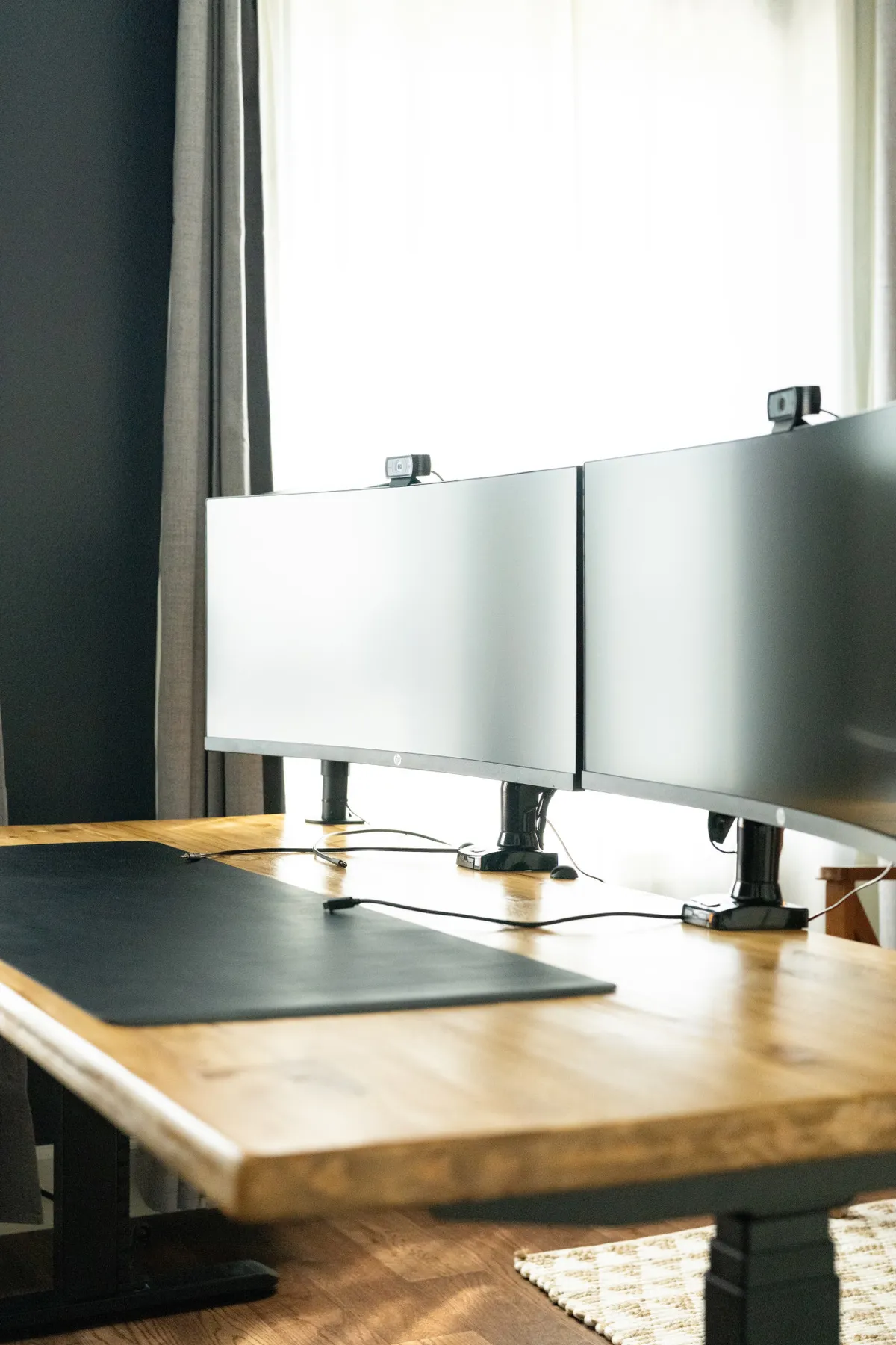 A modern, well-equipped office/music studio with a large wooden desk, dual curved monitors, and a sleek black desk mat. The clean lines and ample workspace convey a productive yet comfortable environment for work, video conferencing, or creative projects.