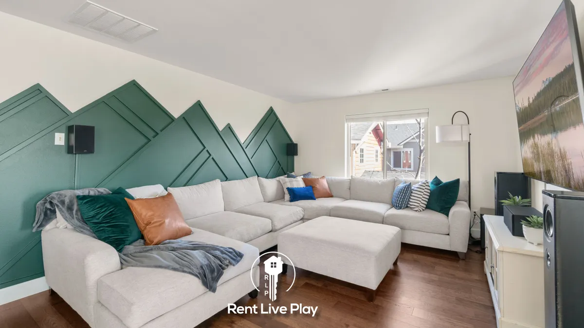 The image showcases a stylish, inviting living room with a bold green accent wall, a plush sectional sofa perfect for gathering, and a large window flooding the space with natural light. The layout encourages relaxation, socializing, and cozy movie nights, perfectly aligning with the property's entertainment amenities.
